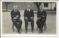 [Group portrait of three men sitting on chairs in an unknown location]