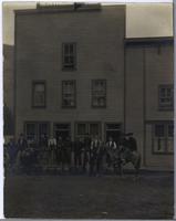 [Group portrait of various men and one horse in front of a storefront in an unknown location]