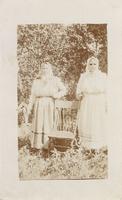 [Portrait of two Doukhobor women standing on either side of a chair in an unknown location]