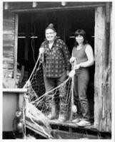 [Man and woman holding fishing nets in the doorway to a wooden building]